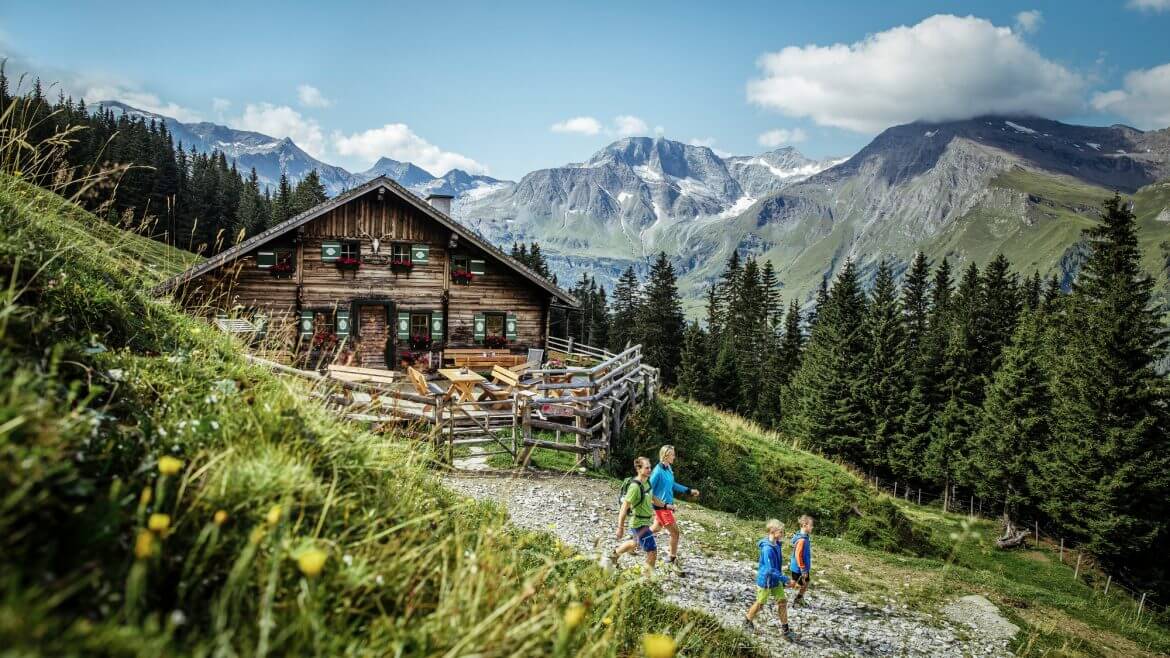Vier Personen wandern auf einen grasbewachsenen Hügel in der Nähe einer Holzhütte mit Bergen und Bäumen im Hintergrund. (vergrößerte Ansicht)