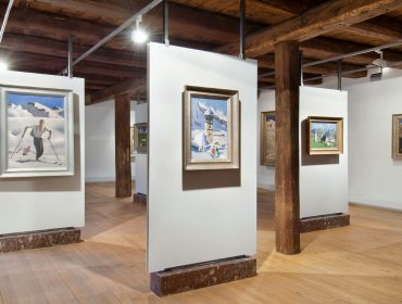 Gemälde mit Gebirgsszenen von Alfons Walde auf weißen Tafeln in einer Galerie mit Holzbalken und Holzfußboden.