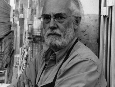 Älterer Mann mit Brille und Bart, in einer Schürze, steht vor einem abstrakten Kunstwerk von Alfons Walde.