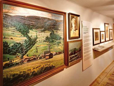 museum-bramberg_stoitzner Gerahmte Landschaftsgemälde und Porträts von Alfons Walde an einer Museumswand mit beschreibenden Texttafeln.
