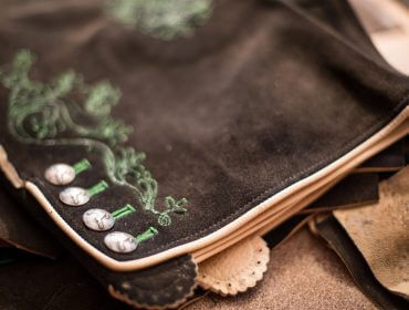 Nahaufnahme einer traditionellen handwerklich bestickten braunen Lederkleidung mit silbernen Knöpfen und grünen Nähten.