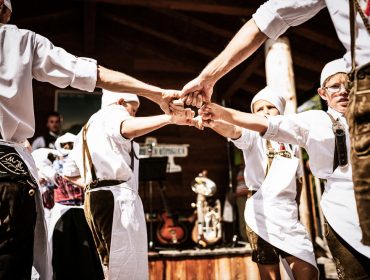 Menschen in traditioneller österreichischer Kleidung halten sich an den Händen und tanzen bei einer Veranstaltung im Freien im Kreis.