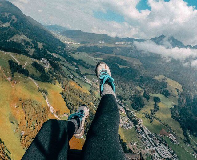 Person beim Gleitschirmfliegen über grünen Hügeln und Tälern, Beine und Schuhe sichtbar, mit Bergen und Wolken in der Ferne.