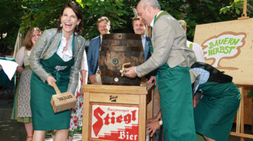 Menschen in Schürzen zapfen bei einer Veranstaltung im Freien ein hölzernes Bierfass an, lächeln und lachen unter grünen Bäumen.