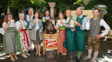 Eine Gruppe von Menschen in traditioneller Kleidung hält Bierkrüge bei einer Stiegl-Bier-Veranstaltung im Freien.