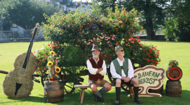 Zwei Männer in österreichischer Tracht sitzen bei einem Blumenwagen mit Bauern Herbst Schild, Fässern und einem großen Heubass.