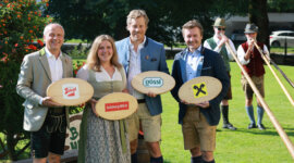 Vier Personen in traditioneller österreichischer Kleidung halten gebrandmarkte Holzbretter, hinter ihnen stehen Alphornbläser auf dem Rasen.