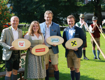 Vier Personen in traditioneller österreichischer Kleidung halten gebrandmarkte Holzbretter, hinter ihnen stehen Alphornbläser auf dem Rasen.