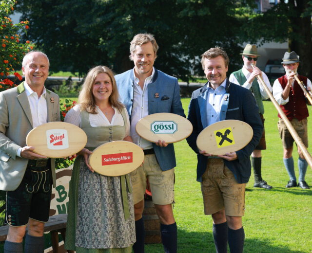Vier Personen in traditioneller österreichischer Kleidung halten gebrandmarkte Holzbretter, hinter ihnen stehen Alphornbläser auf dem Rasen.