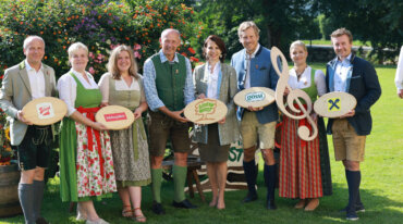 Acht Personen in traditioneller österreichischer Kleidung stehen im Freien, halten verschiedene Holzschilder mit Markenzeichen und lächeln.