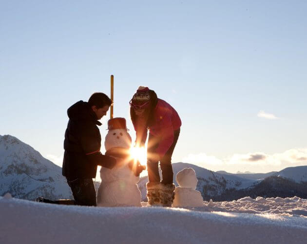 Zwei Personen bauen einen Schneemann auf einem verschneiten Berg bei Sonnenuntergang, durch den das helle Sonnenlicht scheint.