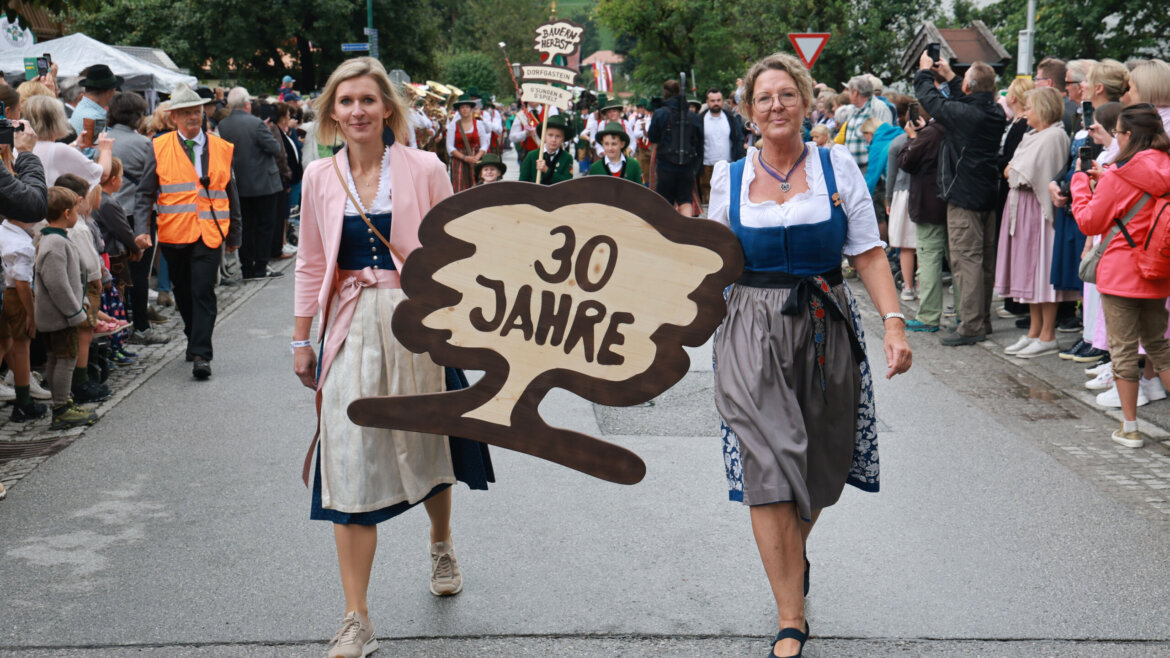 Zwei Frauen in traditioneller Kleidung halten ein 30-Jahre-Schild während einer Straßenparade, umgeben von Menschen. (vergrößerte Ansicht)