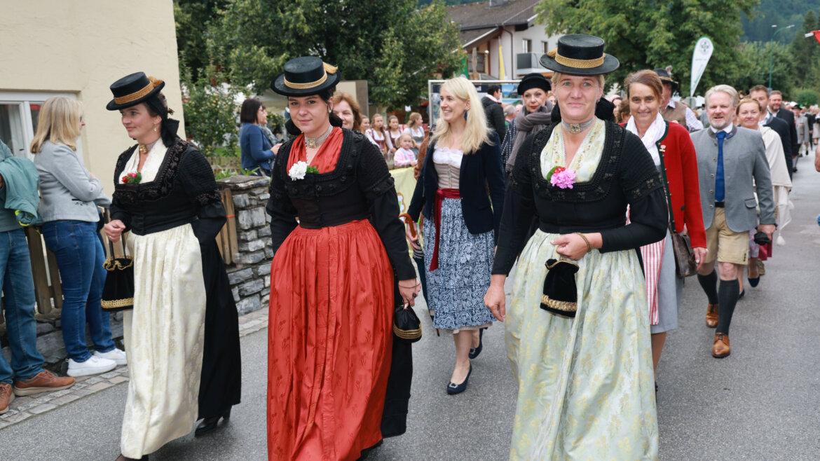 Frauen in österreichischen Trachten und Hüten führen einen Umzug mit festlich gekleideten Menschen durch eine Dorfstraße an. (vergrößerte Ansicht)