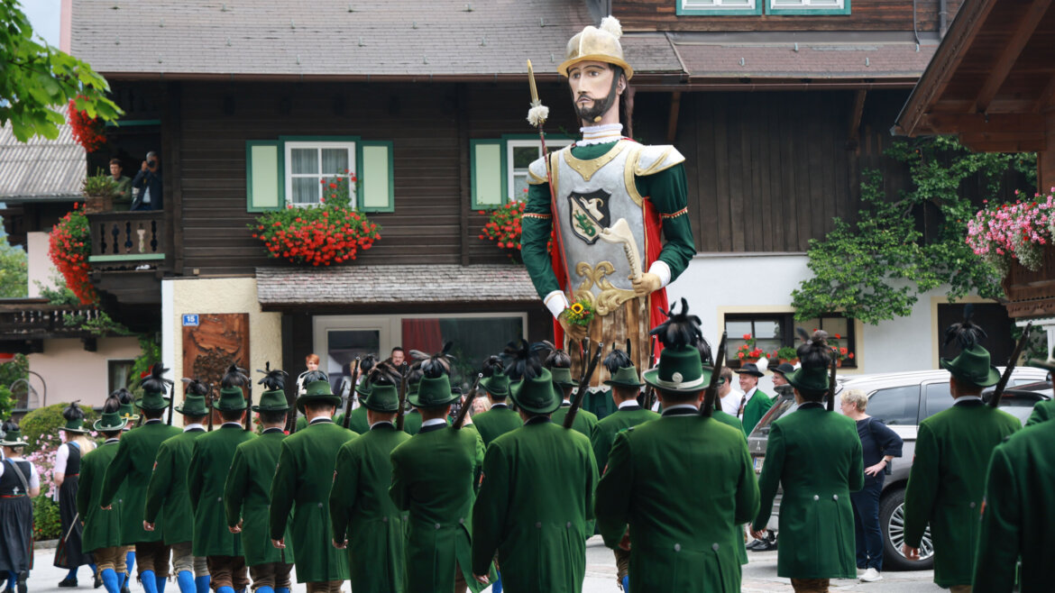 Menschen in grünen Uniformen marschieren auf einen großen Festgiganten in einer traditionellen europäischen Dorfkulisse zu. (vergrößerte Ansicht)