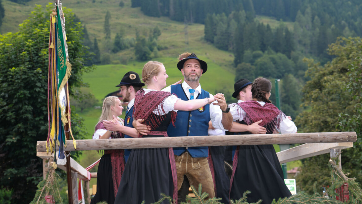 Menschen in österreichischer Tracht tanzen im Freien auf einer Holzbühne mit grünen Hügeln und Bäumen im Hintergrund. (vergrößerte Ansicht)