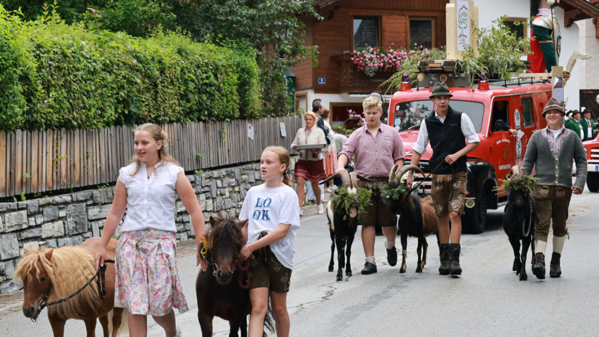 Menschen in traditioneller Kleidung führen Ponys während eines festlichen Umzugs mit Schaulustigen und Oldtimern eine Dorfstraße entlang. (vergrößerte Ansicht)