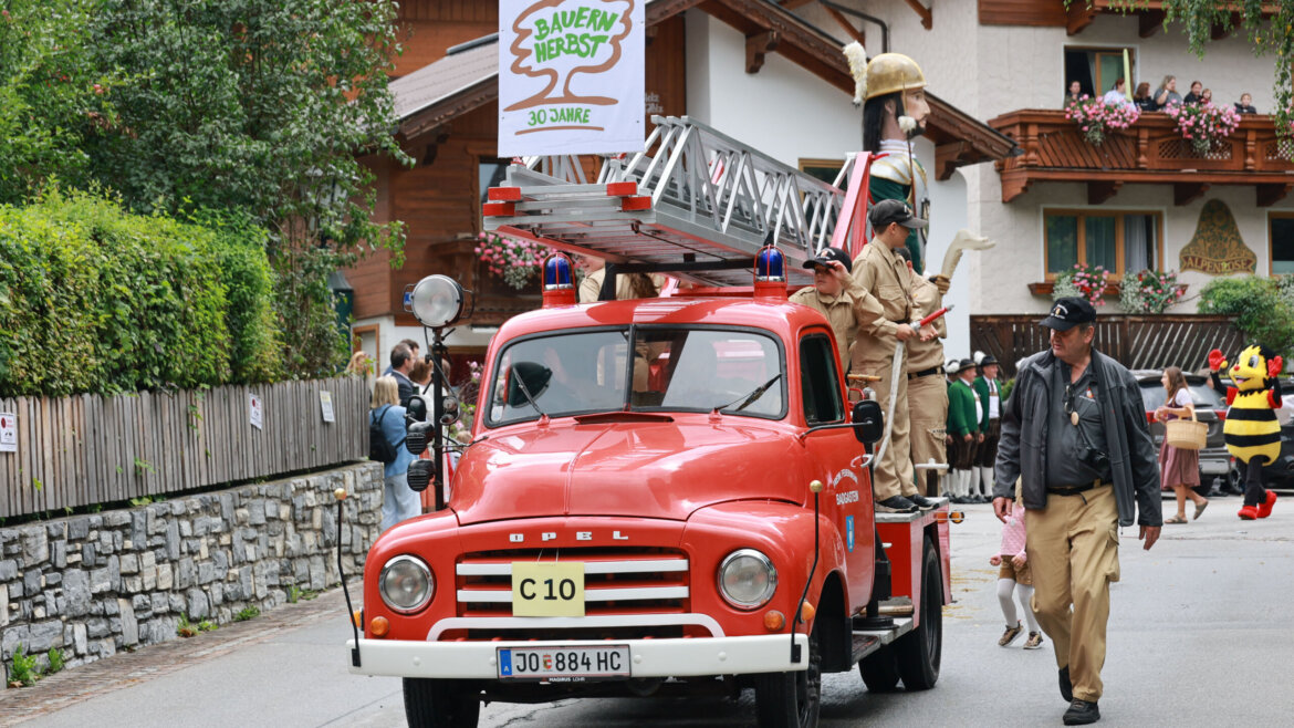 Ein altes rotes Feuerwehrauto in einer Parade, mit Menschen in Uniformen und einem 30 Jahre Bauernherbst-Schild. (vergrößerte Ansicht)