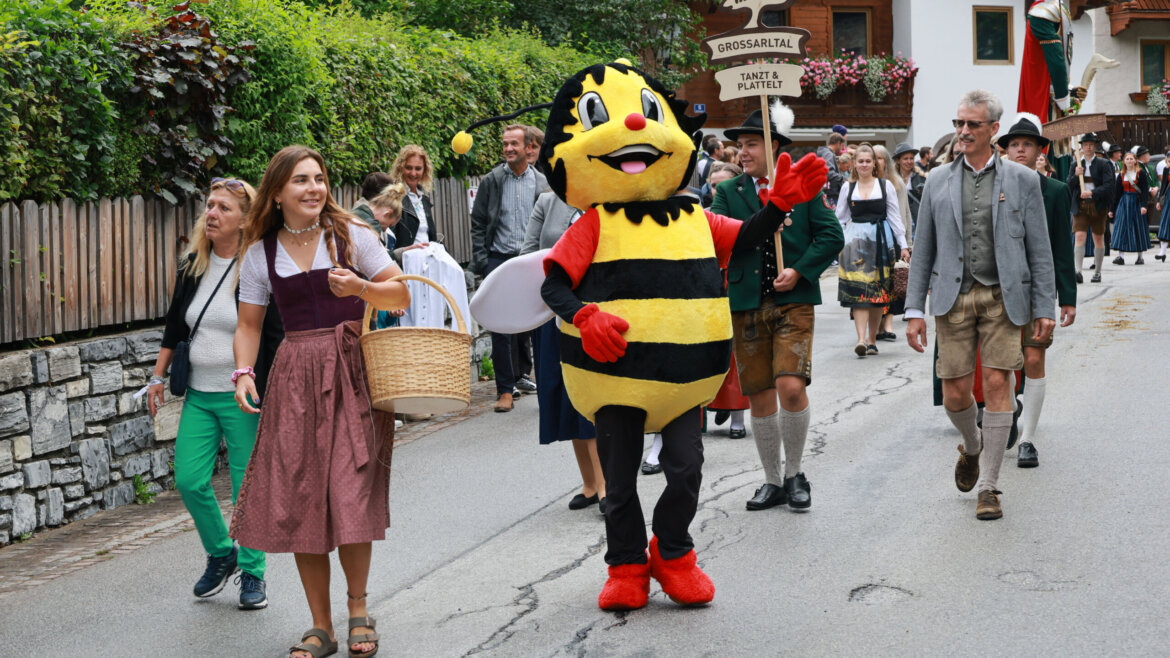 Angeführt von einer Frau mit einem Korb und einer Person im Bienenkostüm ziehen festlich gekleidete Menschen eine Straße entlang. (vergrößerte Ansicht)