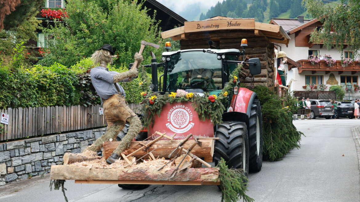 Ein festlicher Traktorumzugswagen mit einer Holzfällerfigur und Baumstämmen, geschmückt mit Blumen und Grünzeug. (vergrößerte Ansicht)
