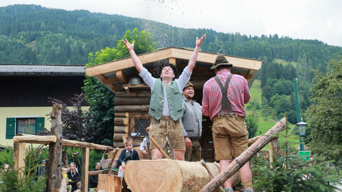 Drei Männer in österreichischer Tracht feiern an einem großen Baumstamm in einem Alpendorf mit Bergkulisse. (vergrößerte Ansicht)
