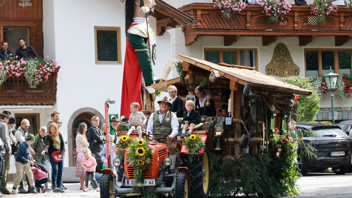 In einem festlichen Umzug ziehen ein Traktor mit Sonnenblumen und Menschen in österreichischer Tracht an einem Gebäude im alpenländischen Stil vorbei. (vergrößerte Ansicht)