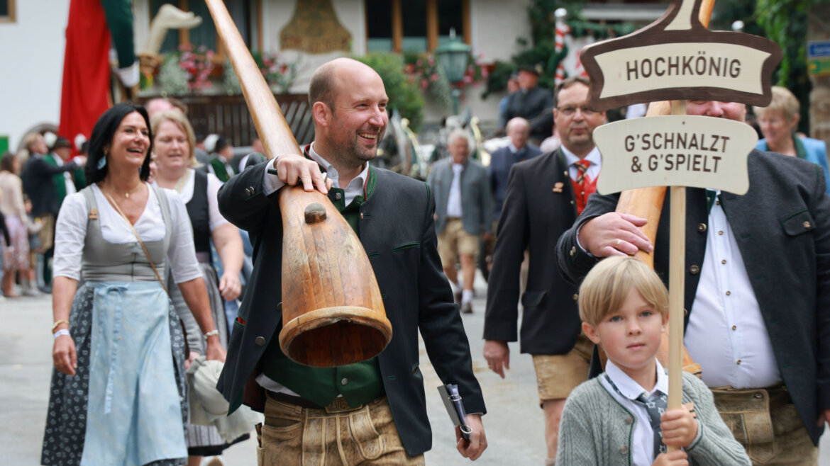 Menschen in traditioneller österreichischer Kleidung laufen in einer Parade; ein Mann trägt ein Alphorn, ein Junge hält ein Schild. (vergrößerte Ansicht)