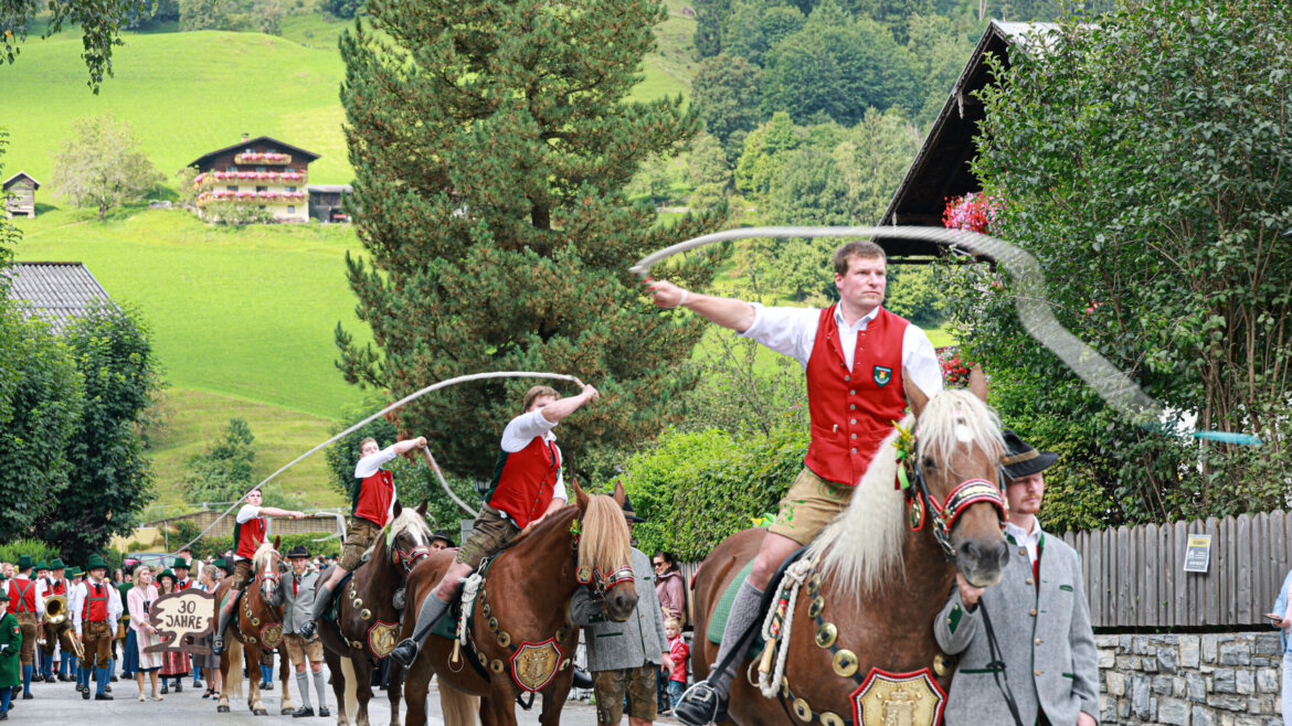 Männer in traditioneller österreichischer Kleidung reiten auf Pferden und lassen Peitschen knallen bei einem festlichen Umzug durch ein ländliches, bergiges Dorf. (vergrößerte Ansicht)