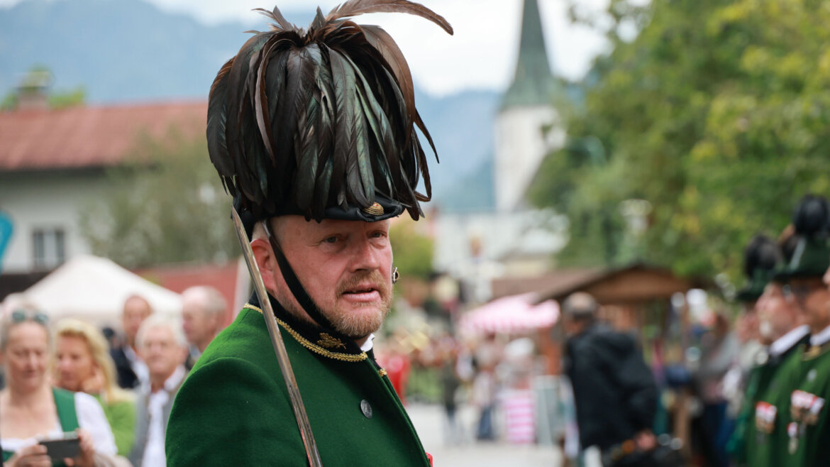 Mann in traditioneller grüner Kleidung mit einem großen Federhut bei einem Fest im Freien, Menschen im Hintergrund. (vergrößerte Ansicht)