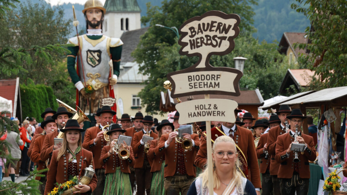 Eine Frau führt eine traditionelle Marschmusikparade mit einem Schild in einer festlichen Dorfumgebung an. (vergrößerte Ansicht)