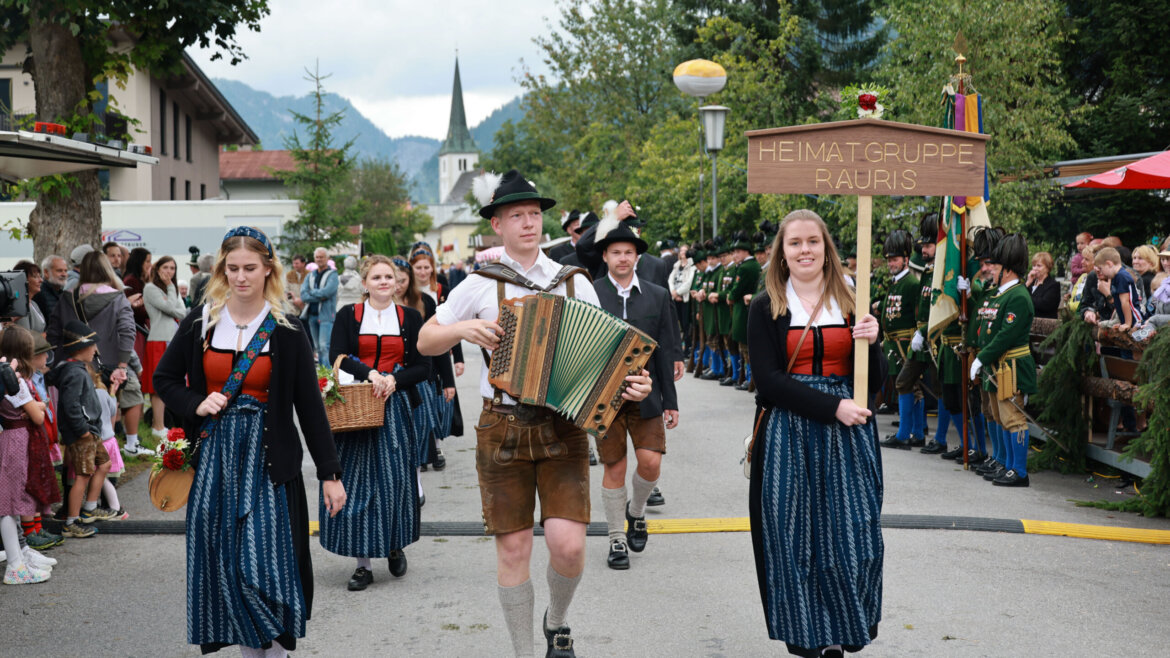 Menschen in traditioneller österreichischer Kleidung ziehen mit Musikern und einem Schild mit der Aufschrift "Heimatgruppe Rauris" umher. (vergrößerte Ansicht)