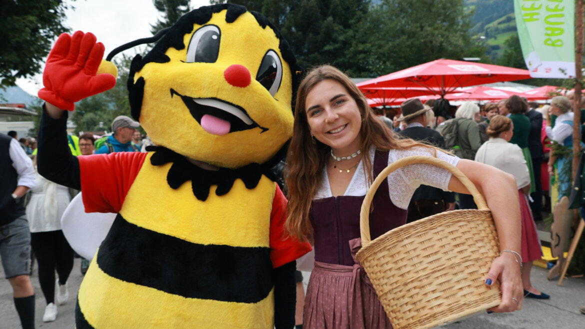 Eine Person in einem Bienenkostüm steht neben einer lächelnden Frau in österreichischer Tracht, die bei einer Veranstaltung im Freien einen Korb hält. (vergrößerte Ansicht)