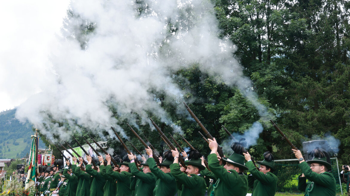 Eine Reihe von Personen in grünen Uniformen schießt mit Gewehren in die Luft und erzeugt weiße Rauchwolken im Freien. (vergrößerte Ansicht)