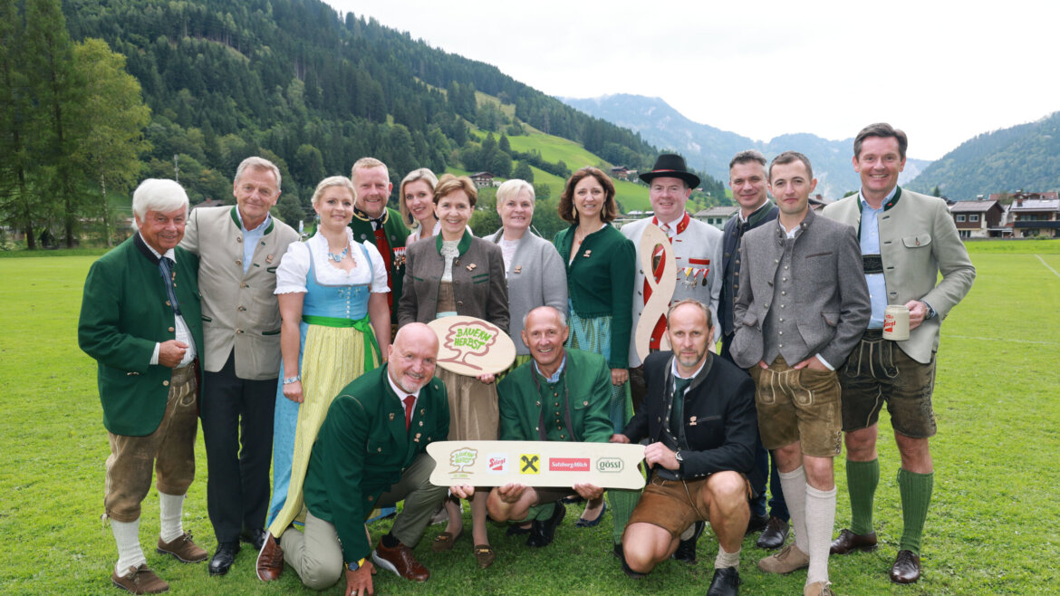 Eine Gruppe von Menschen in traditioneller österreichischer Kleidung steht im Freien auf einer Wiese mit den Bergen im Hintergrund zusammen. (vergrößerte Ansicht)