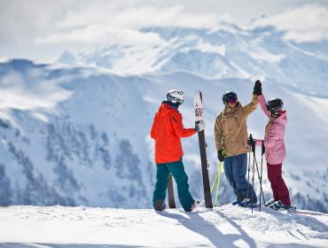Drei Skifahrer in Winterausrüstung geben sich auf einem verschneiten Skicircus-Berg die Hand, mit schneebedeckten Gipfeln hinter ihnen.
