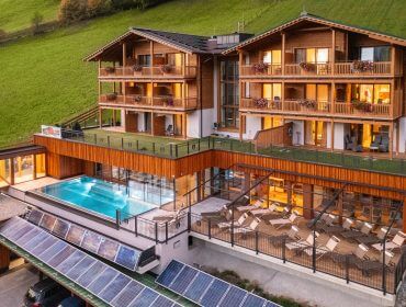 Modernes Holzhotel mit Balkonen, Außenpool, Sonnenliegen und einem Berghang im Hintergrund bei Sonnenuntergang.
