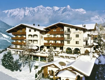 Ein großes Alpenhotel, umgeben von Schnee, mit Bergen im Hintergrund und blauem Himmel.