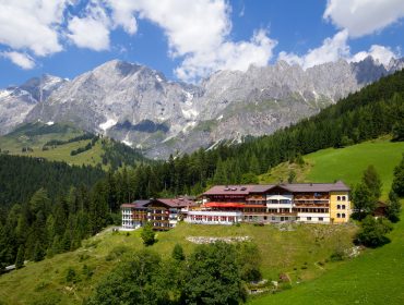 Hotelgebäude an einem grünen Hang mit hohen Bergen und blauem Himmel im Hintergrund.