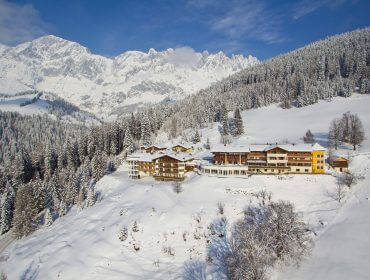Ein großes Hotel liegt an einem verschneiten Hang, umgeben von Kiefern und Bergen unter blauem Himmel.