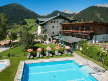 Alpenhotel mit Pool, Liegestühlen und Blick auf die Berge unter einem strahlend blauen Himmel.