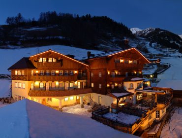 Ein großes, warm beleuchtetes Hotel im österreichischen Chalet-Stil, umgeben von Schnee und Bergen in der Abenddämmerung.
