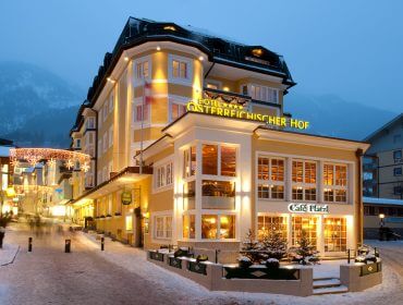 Ein warm beleuchtetes Hotel und ein Café stehen auf einer verschneiten Straße in einer winterlichen Bergstadt in der Abenddämmerung.