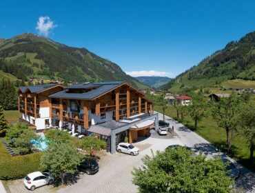 Ein großes alpines Hotel mit hölzernen Akzenten, eingebettet in ein üppig grünes Bergtal unter einem klaren blauen Himmel.