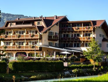 Ein großes Hotel im alpinen Stil mit Balkonen und Blumenkästen, umgeben von viel Grün und Bergen.