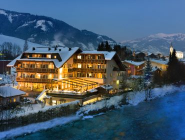 Ein warm beleuchtetes Hotel in einem verschneiten österreichischen Bergdorf am Ufer eines Flusses in der Abenddämmerung.