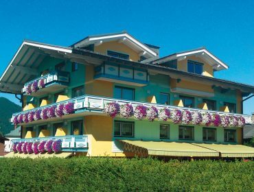 Ein gelbes Haus mit Balkonen, die mit lila und weißen Blumen geschmückt sind, unter einem klaren blauen Himmel.