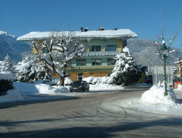 Ein schneebedecktes Haus mit einem geparkten Auto davor, umgeben von verschneiten Bäumen und Bergen im Hintergrund.