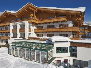 Ein hölzernes Alpenhotel mit verschneiten Dächern und einer gläsernen Sonnenterrasse, umgeben von schneebedecktem Boden unter blauem Himmel.