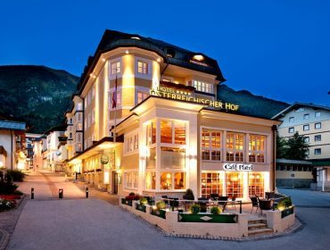 Ein hell erleuchtetes Hotel mit Sitzplätzen im Freien an einer ruhigen Straße in der Abenddämmerung, umgeben von Bergen.