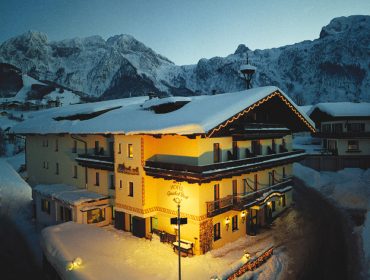 Verschneites Alpenhotel in der Abenddämmerung mit leuchtenden Lichtern, umgeben von Bergen und tiefem Winterschnee.