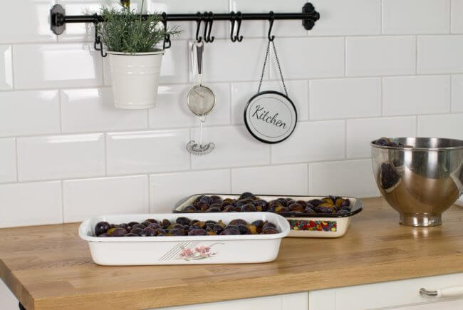 Zwei Tabletts und eine mit Pflaumen gefüllte Metallschüssel auf einer hölzernen Küchentheke mit weiß gefliester Wand.